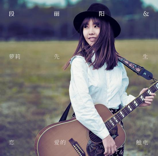 写真中的段丽阳头戴黑色毡帽,手中抱着一把木吉他,站在郊外的草地上